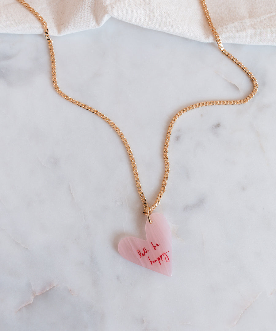 The WALD Berlin "Let's be happy" gold necklace features an 18k gold-plated chain with a pink heart-shaped hand-painted pendant inscribed with "don't be happy," stylishly displayed on a marble surface.