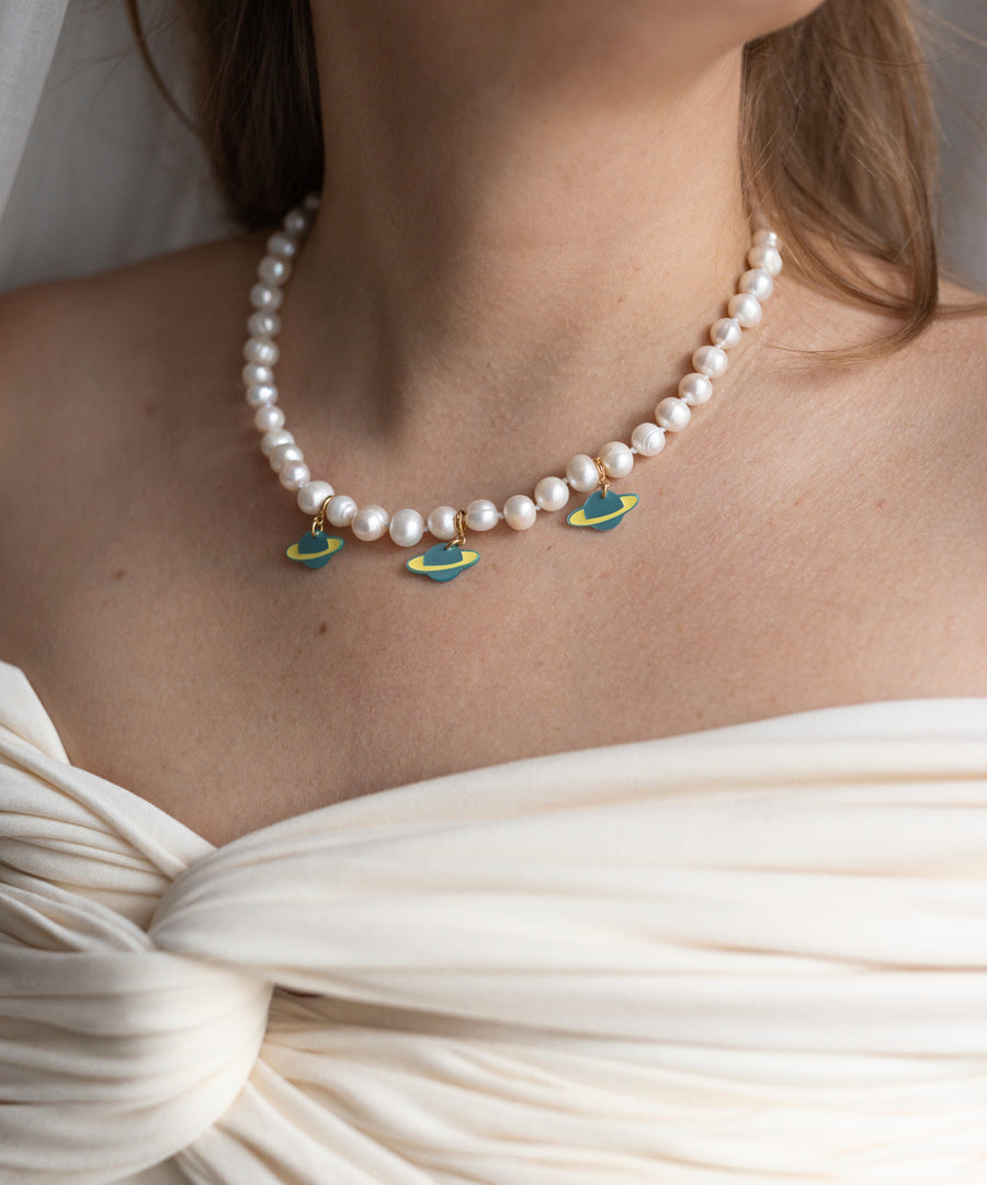 A woman wears the WALD Berlin SAMPLE SALE From Another Mother Galactic Pearl Necklace, featuring sweetwater pearls and three Saturn charms set in 925 sterling silver, paired with an off-shoulder white dress.