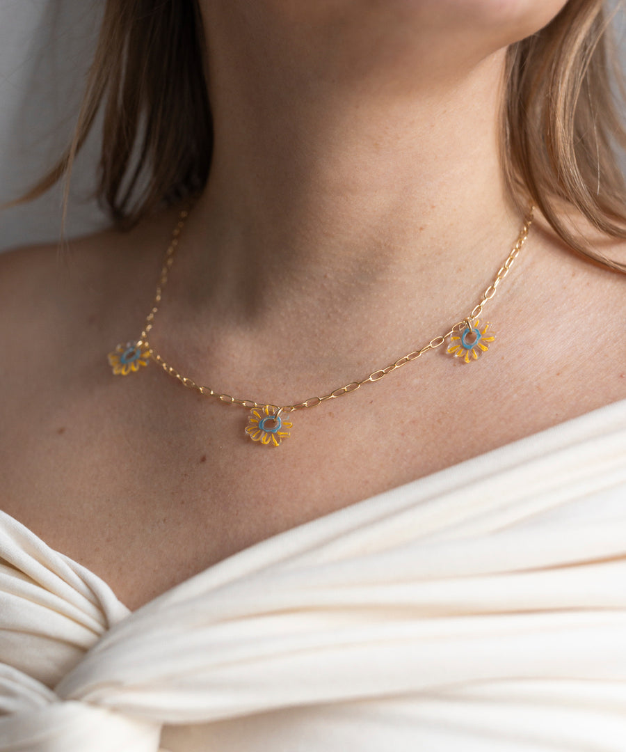 The SAMPLE SALE Daisy How High Necklace by WALD Berlin features three blue and yellow daisy charms on an 18k gold-plated chain—shown with an off-white draped top. A bright, everyday piece.