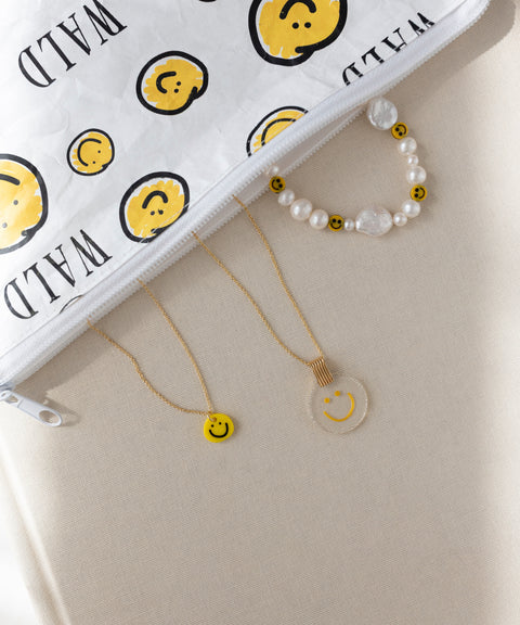 Eine weiße Reißverschlusstasche mit gelben Smiley-Motiven, ein WALD Berlin Smilie Dude Perlenarmband mit Verlängerung, eine runde Smiley-Halskette und eine gelbe Smiley-Halskette.