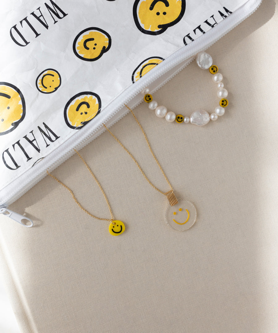 Eine weiße Reißverschlusstasche mit gelben Smiley-Motiven, ein WALD Berlin Smilie Dude Perlenarmband mit Verlängerung, eine runde Smiley-Halskette und eine gelbe Smiley-Halskette.