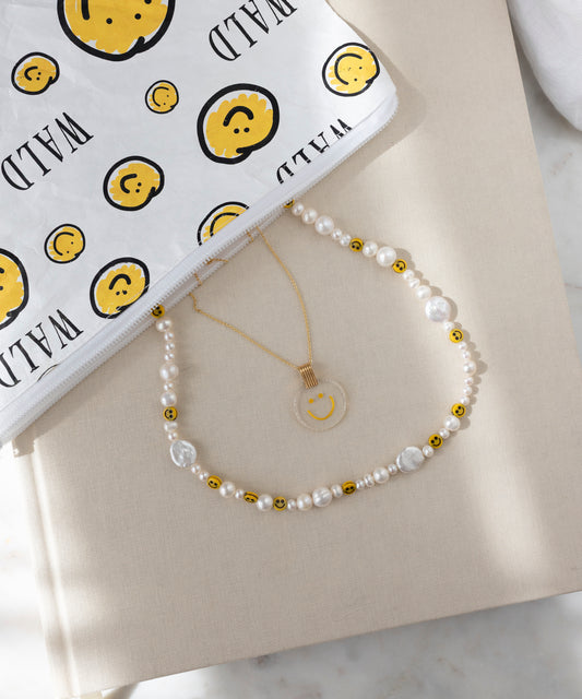 The WALD Berlin Smilie Dude Pearl Necklace, handmade with sweetwater pearls, gold beads, and smiley faces, is displayed beside a gold pendant necklace and a white pouch with yellow smileys and "WILD" text.
