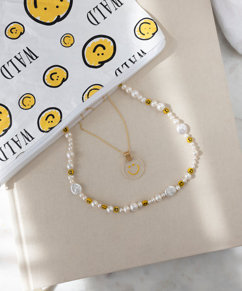 Die Smilie Dude Medaillon Gold Halskette von WALD Berlin besteht aus einem klaren Smiley-Anhänger an einer vergoldeten Kette, der neben einem weißen "WILD"-Beutel mit Smiley-Gesicht auf Beige steht. Hergestellt in Deutschland.