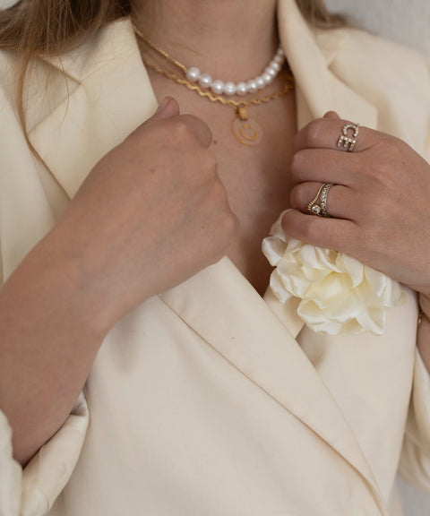 Eine Person trägt einen cremefarbenen Blazer mit einer weißen Reversblume, eine Perlenkette, eine WALD Berlin Irina Halskette Gold, Ringe an der linken Hand und ein Messingarmband.