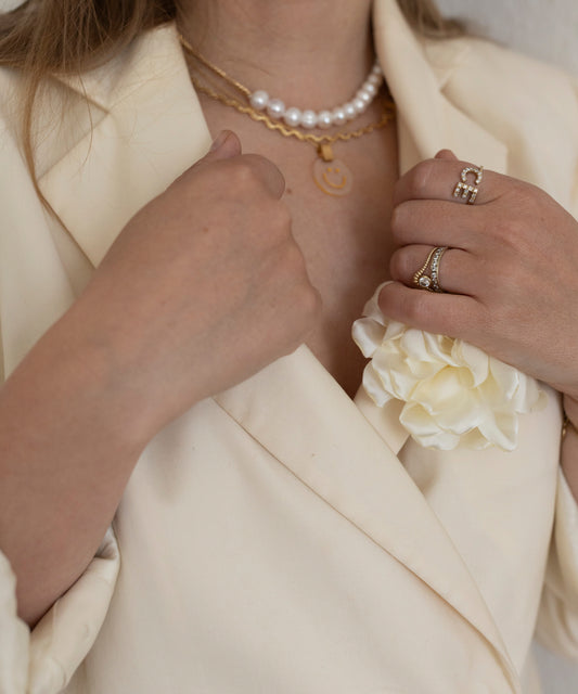 Eine Person trägt einen cremefarbenen Blazer mit einer weißen Reversblume, eine Perlenkette, eine WALD Berlin Irina Halskette Gold, Ringe an der linken Hand und ein Messingarmband.