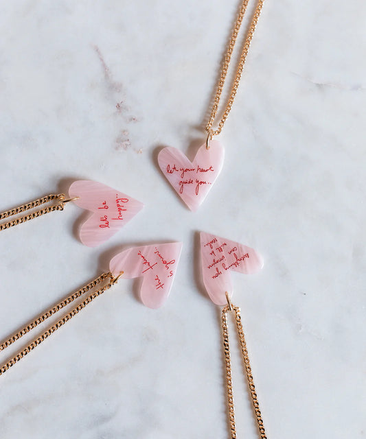 Vier rosafarbene herzförmige WALD Berlin "Let Your Heart Guide You"-Anhänger mit vergoldeten Ketten sind kreisförmig auf Marmor angeordnet und jeweils mit roter Kursivschrift graviert - eine ideale Halskette mit Herz oder ein persönliches Geschenk.