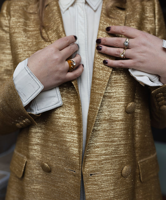A person in a shiny gold blazer made from bio polyester, over a white shirt, showcases Isla Bonita shining star rings by WALD Berlin adorned with Swarovski stones and wears dark nail polish.