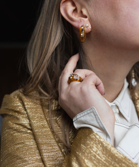 A person wearing a gold-textured blazer and white shirt showcases hoop earrings and WALD Berlin's Isla Bonita shining star ring with amber-colored Swarovski stones.