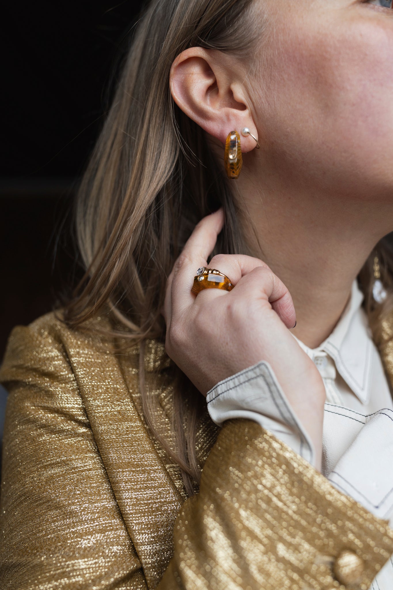 A person wearing a gold-textured blazer and white shirt showcases hoop earrings and WALD Berlin's Isla Bonita shining star ring with amber-colored Swarovski stones.