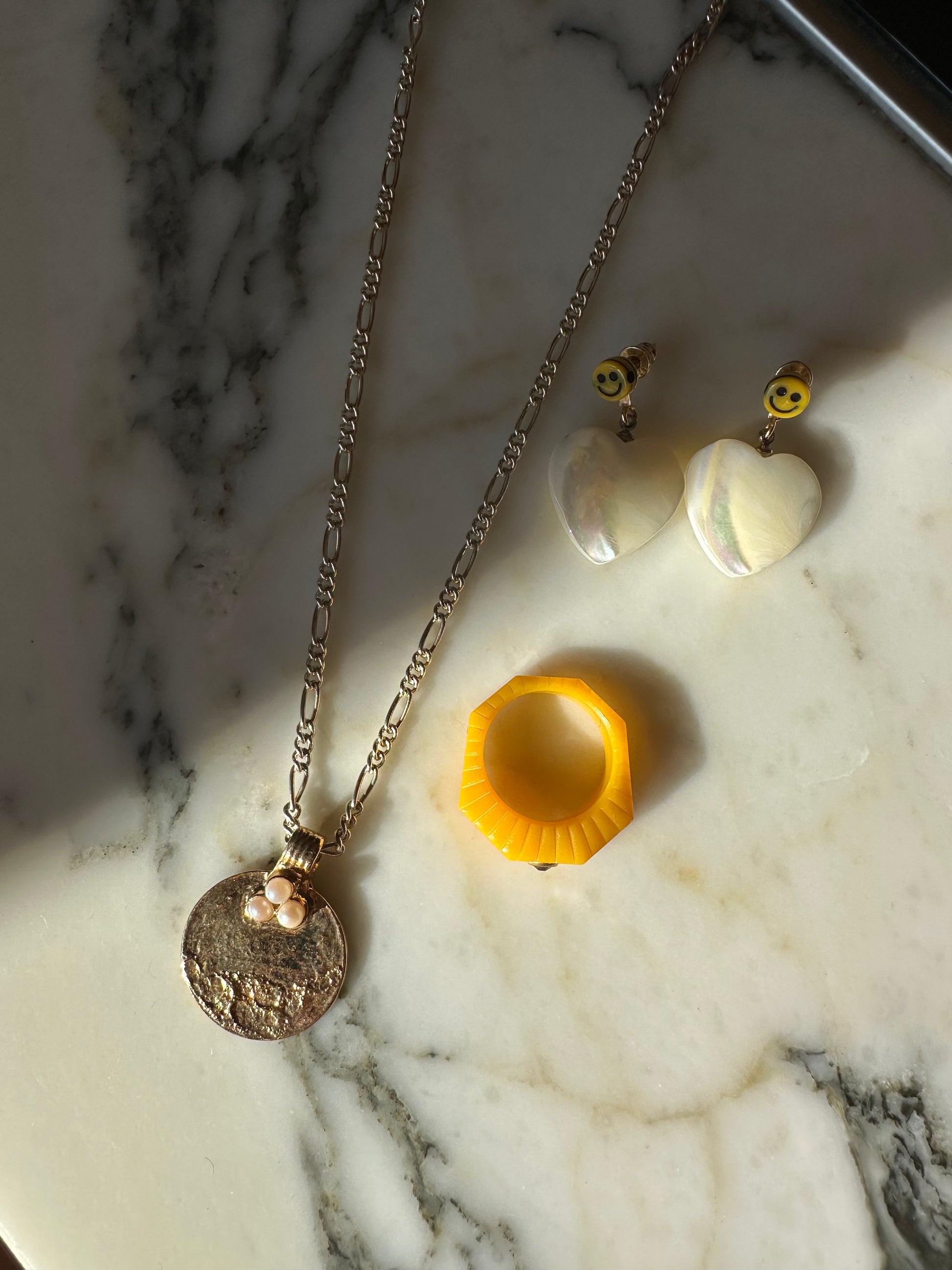A gold necklace with a charming pendant, a yellow ring, and heart-shaped earrings adorned with smiley faces rest elegantly on the marble surface, paired with a chic WALD Berlin surprise bag.