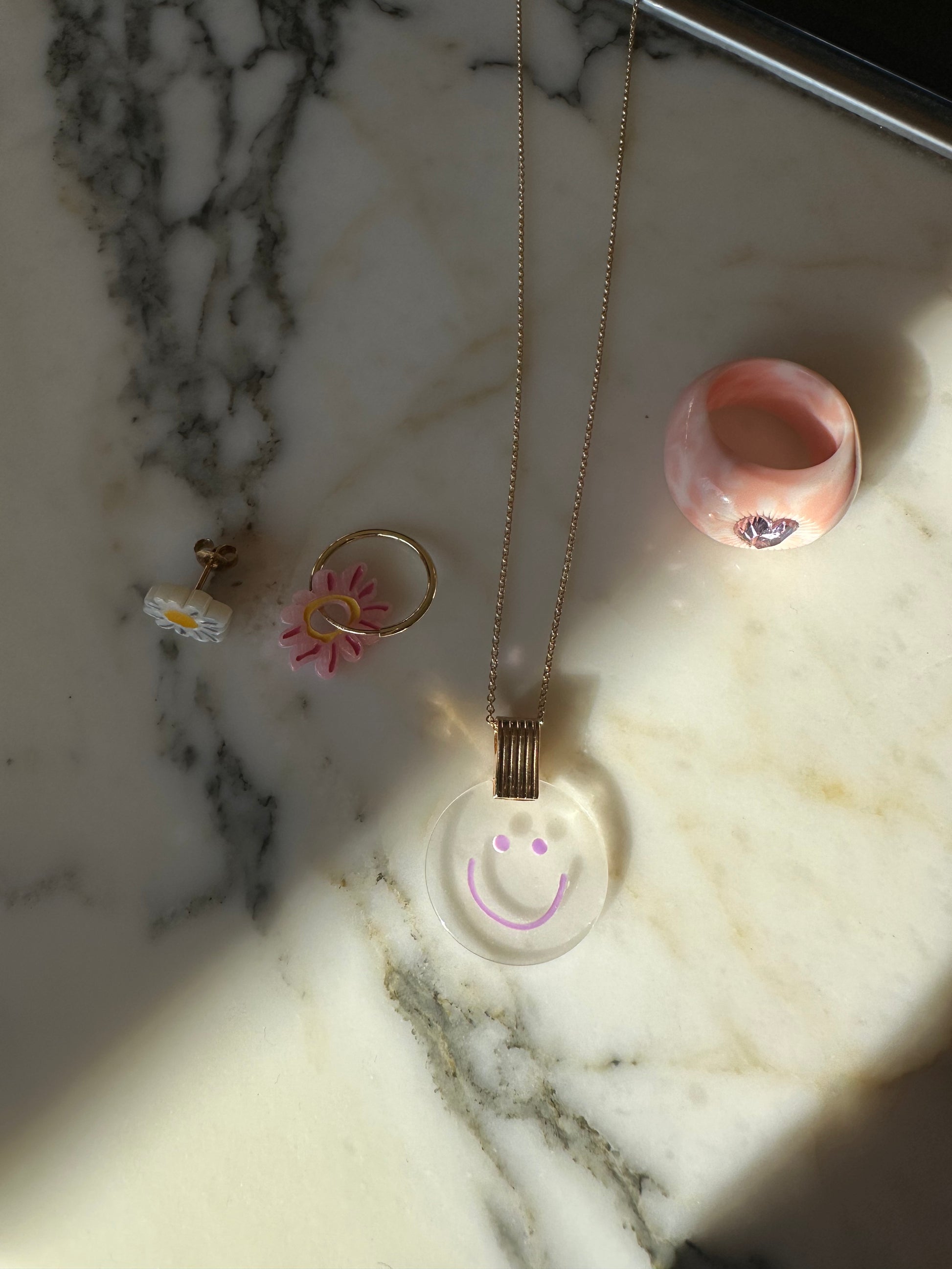 A smiley face necklace, pink flower ring, white flower earrings, and a pink marble-like ring are elegantly displayed on a marble surface, echoing the chic simplicity of a surprise WALD Berlin Bag by WALD Berlin.