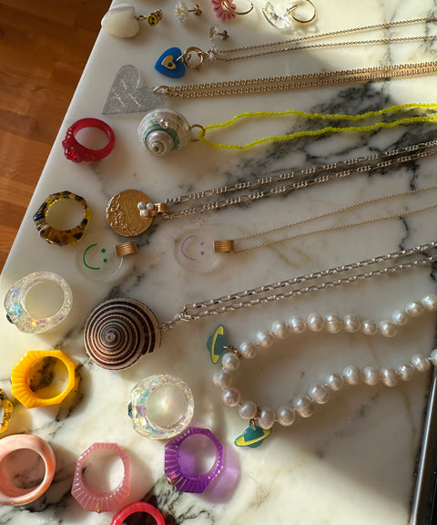 A variety of necklaces, rings, and earrings are beautifully displayed on marble. This collection artfully blends materials like pearls, beads, and metals. The vibrant colors and playful designs enhance the chic vibe of a surprise WALD Berlin Bag by WALD Berlin.