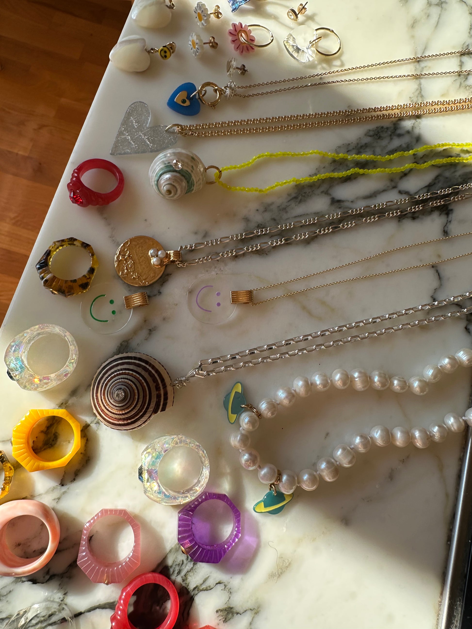 A variety of necklaces, rings, and earrings are beautifully displayed on marble. This collection artfully blends materials like pearls, beads, and metals. The vibrant colors and playful designs enhance the chic vibe of a surprise WALD Berlin Bag by WALD Berlin.