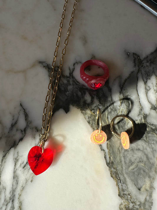 A necklace with a red heart pendant and a pink ring is arranged beside two gold hoop earrings with pink smiley face charms on marble, echoing the playful elegance of a surprise WALD Berlin Bag by WALD Berlin.