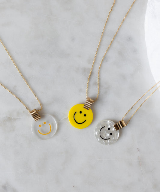 Three smiley face pendants in spring colors: a WALD Berlin Smilie Dude Medaillon Pearl Necklace, one clear, one yellow, and one pearlescent, shimmer on a marble surface—each 18k gold-plated for elegance.