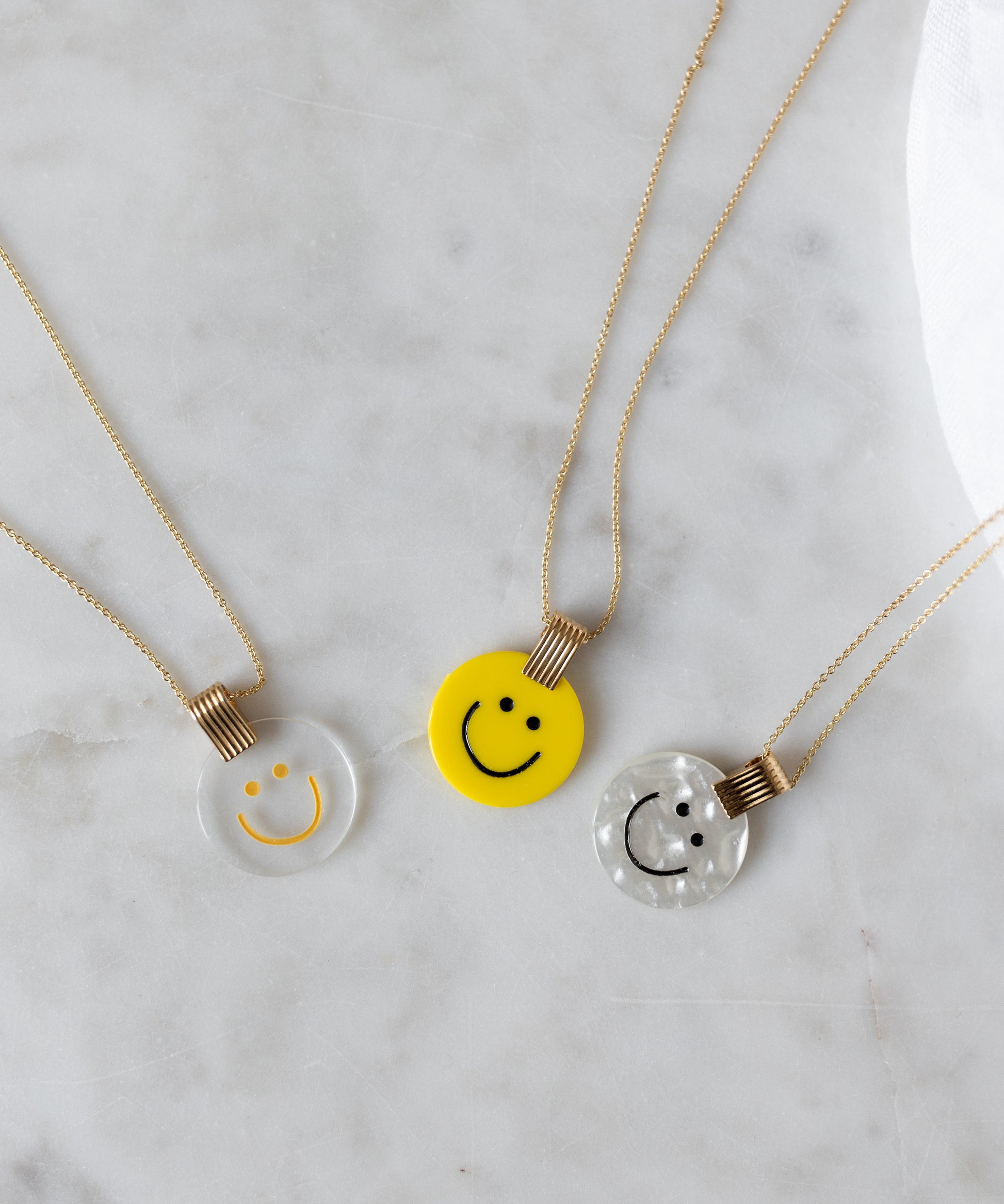Three smiley face pendants in spring colors: a WALD Berlin Smilie Dude Medaillon Pearl Necklace, one clear, one yellow, and one pearlescent, shimmer on a marble surface—each 18k gold-plated for elegance.
