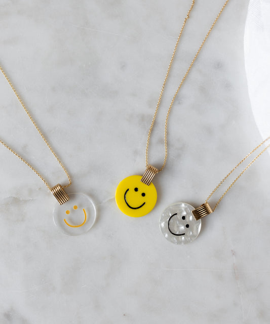 Three smiley face pendants in spring colors: a WALD Berlin Smilie Dude Medaillon Pearl Necklace, one clear, one yellow, and one pearlescent, shimmer on a marble surface—each 18k gold-plated for elegance.