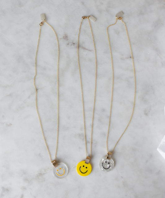 Three WALD Berlin Smilie Dude Medaillon Yellow Necklaces with hand-painted polyester and 18k gold-plated brass pendants in clear, yellow, and light gray rest on a white marble surface.