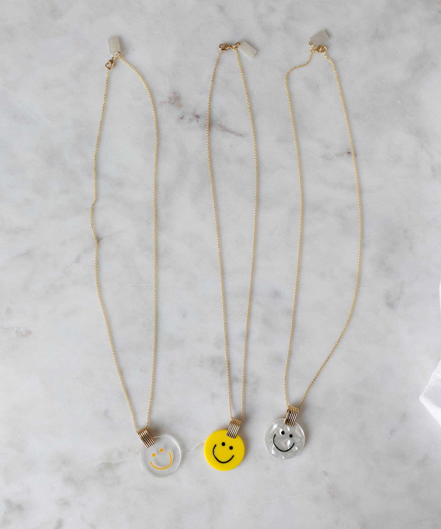 Three WALD Berlin Smilie Dude Medaillon Yellow Necklaces with hand-painted polyester and 18k gold-plated brass pendants in clear, yellow, and light gray rest on a white marble surface.