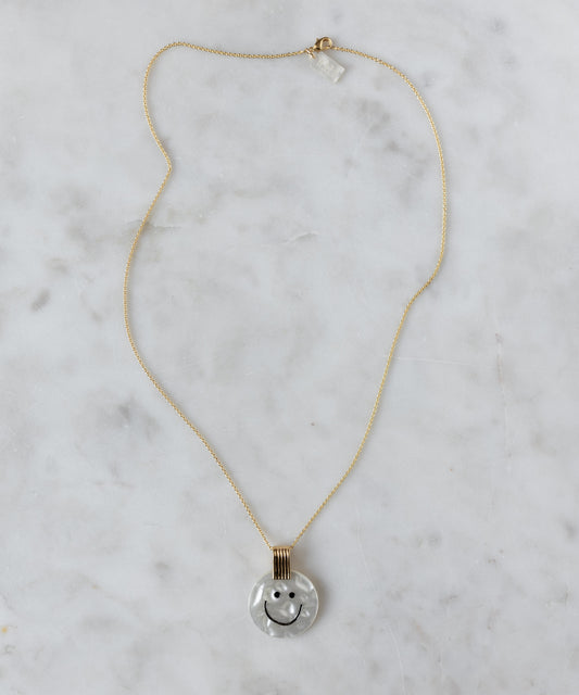 The WALD Berlin Smilie Dude Medaillon Pearl Necklace features an 18k gold-plated chain and round pendant with a smiley face on a white marbled background, adding cheerful spring colors to any outfit.