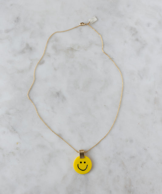 The WALD Berlin Smilie Dude Medaillon Yellow Necklace, featuring an 18k gold-plated brass chain and a yellow smiley face pendant, rests elegantly on a marbled surface.