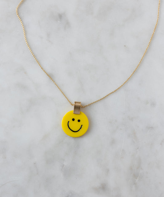 The WALD Berlin Smilie Dude Medaillon Yellow Necklace boasts an 18k gold-plated brass chain with a cheerful yellow smiley face pendant, elegantly displayed on a sleek marble surface.