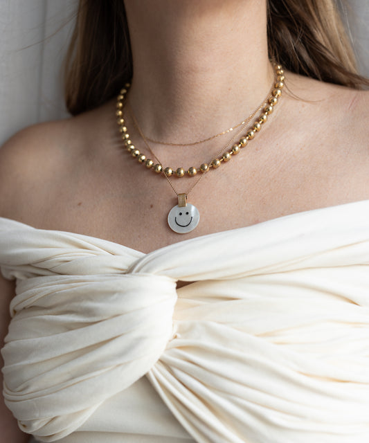 A woman wearing an off-shoulder white top showcases the WALD Berlin Smilie Dude Medaillon Pearl Necklace, featuring 18k gold-plated beads that reflect spring colors.