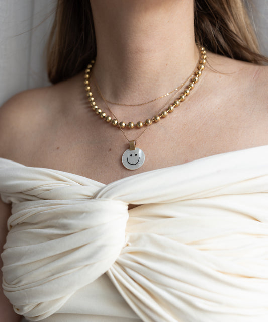 A woman wearing an off-shoulder white top showcases the WALD Berlin Smilie Dude Medaillon Pearl Necklace, featuring 18k gold-plated beads that reflect spring colors.