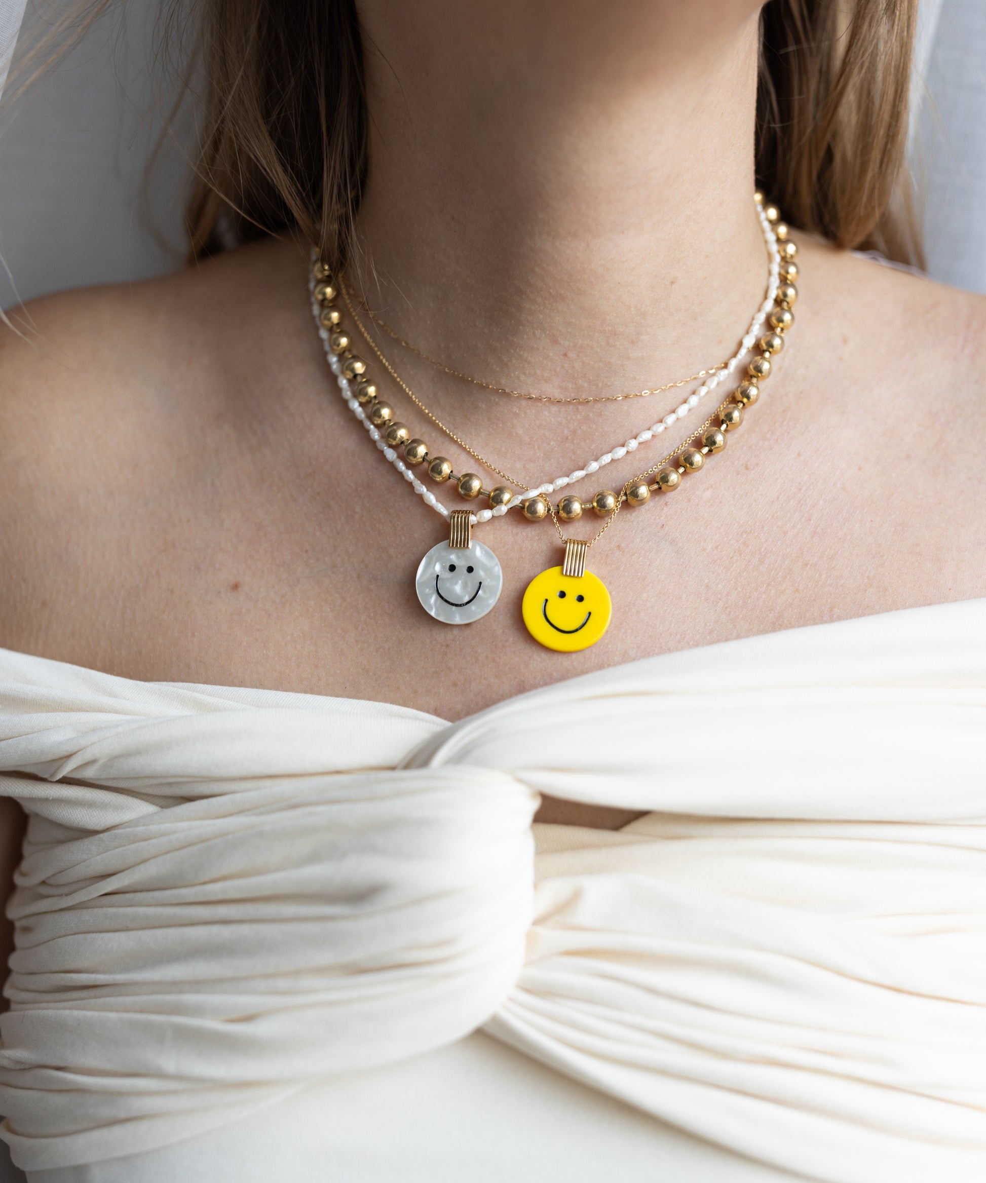 A woman shines in layered necklaces, featuring two metallic and one WALD Berlin Smilie Dude Medaillon Yellow Necklace, all 18k gold-plated brass, elegantly draped over an off-the-shoulder white top.