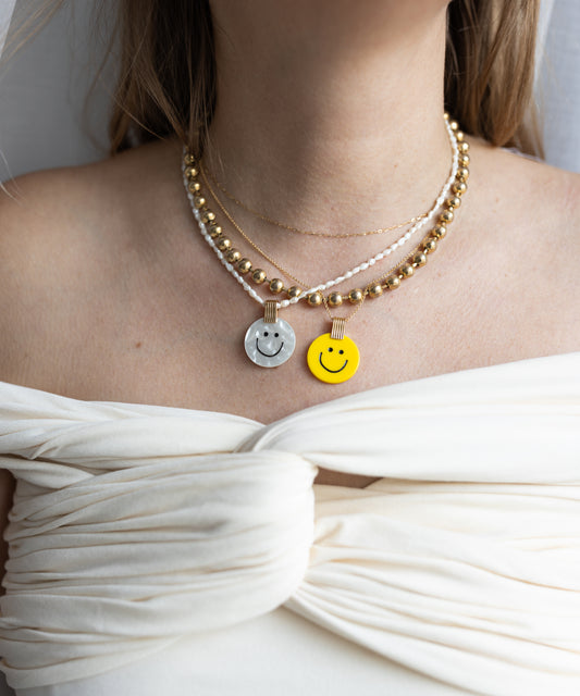 A woman shines in layered necklaces, featuring two metallic and one WALD Berlin Smilie Dude Medaillon Yellow Necklace, all 18k gold-plated brass, elegantly draped over an off-the-shoulder white top.