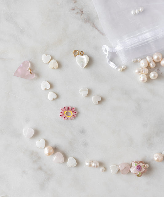Assorted heart-shaped beads, a pink flower bead, and unique pearls from the Sweet Water Pearls Kit by WALD World are arranged on a marble surface. A sheer white drawstring pouch and more pearls from the DIY collection can be seen in the background.