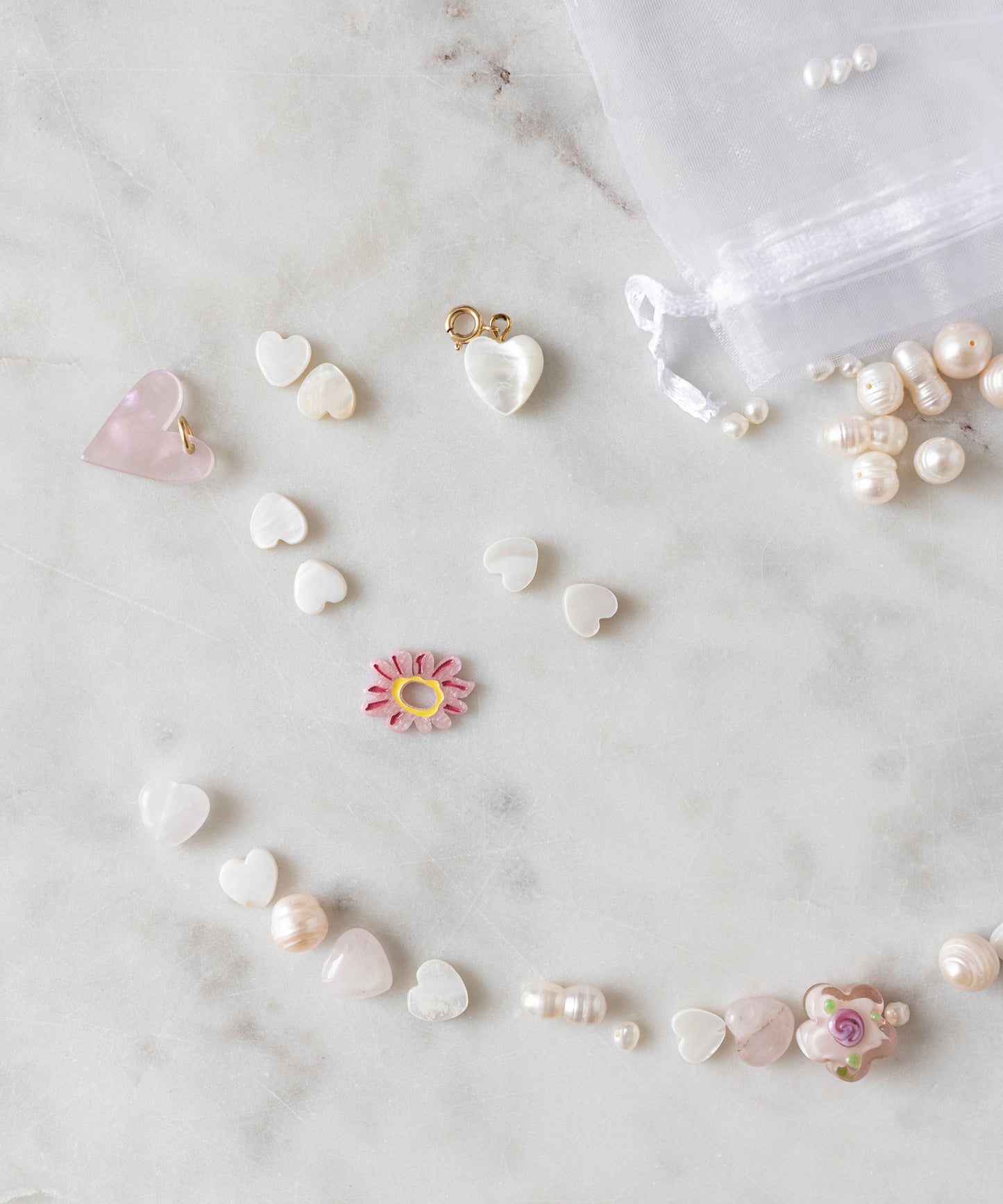 Assorted heart-shaped beads, a pink flower bead, and unique pearls from the Sweet Water Pearls Kit by WALD World are arranged on a marble surface. A sheer white drawstring pouch and more pearls from the DIY collection can be seen in the background.