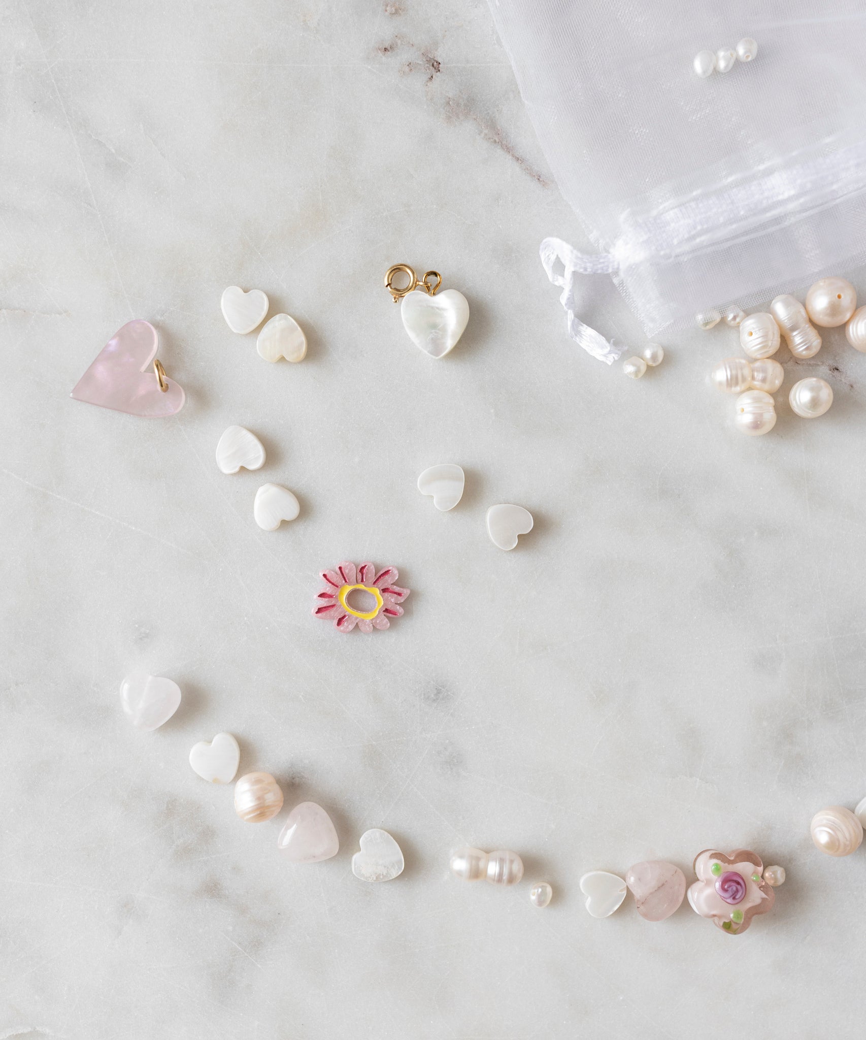A scattering of various Daisy Charm Rose beads, pearls, and charms from WALD World on a white marble surface next to a small, translucent drawstring bag with some pearls spilling out invites opportunities for creative crafting and DIY jewelry projects.