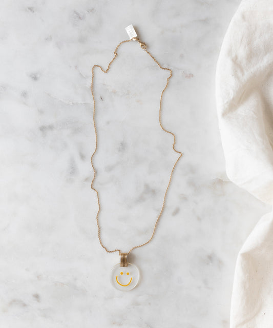 The Smilie Dude Medaillon Gold Necklace by WALD Berlin gleams delightfully on the marble surface, showcasing its exquisite craftsmanship. Made in Germany, this 18k gold-plated accessory combines cheerful elegance with impeccable quality.