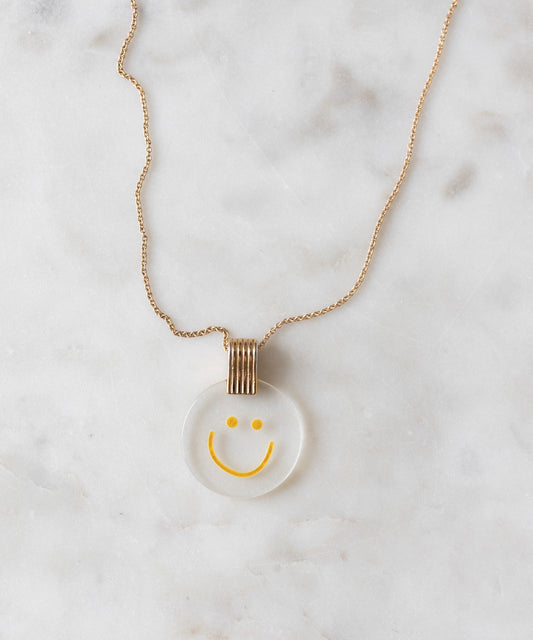 The Smilie Dude Medaillon Gold Necklace from WALD Berlin features an 18k gold-plated chain adorned with a round smiley medallion pendant against a white marble backdrop, crafted with precision and elegance. Made in Germany, it beautifully combines playful charm with sophisticated artistry.