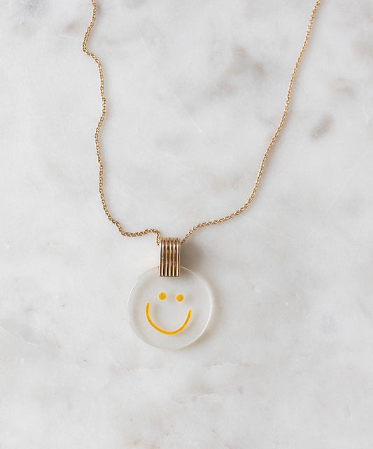 The Smilie Dude Medaillon Gold Necklace from WALD Berlin features an 18k gold-plated chain adorned with a round smiley medallion pendant against a white marble backdrop, crafted with precision and elegance. Made in Germany, it beautifully combines playful charm with sophisticated artistry.