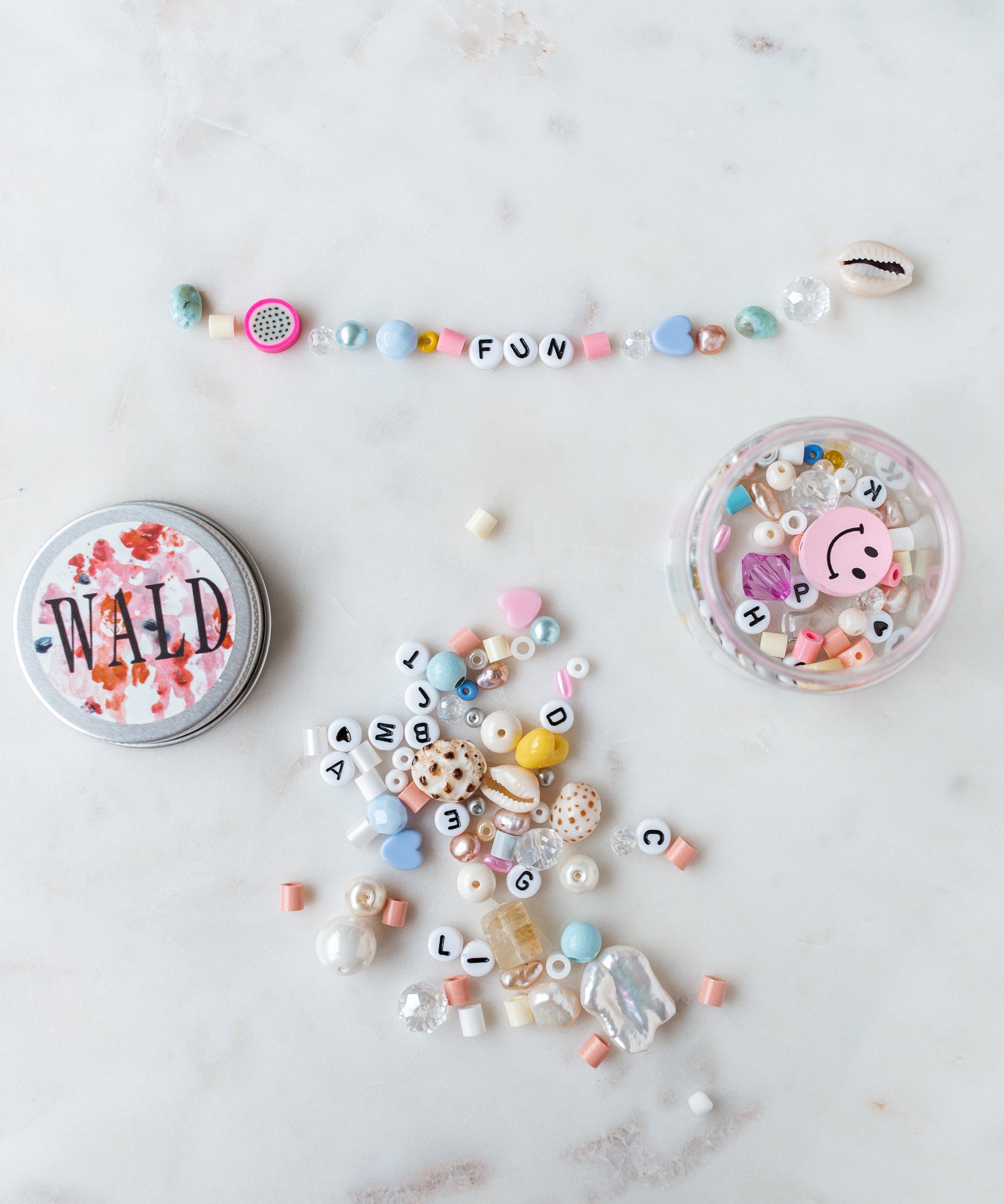 Assorted beads and a bracelet spelling out 'FUN' are arranged on a marble surface. Nearby, there is a tin labeled "WILD" and a DIY box with a smiley face, filled with more beads from the WALD World Candyman Kit in powder candy tones.