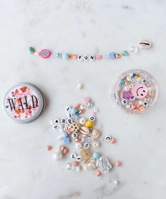 Assorted beads and a bracelet spelling out 'FUN' are arranged on a marble surface. Nearby, there is a tin labeled "WILD" and a DIY box with a smiley face, filled with more beads from the WALD World Candyman Kit in powder candy tones.