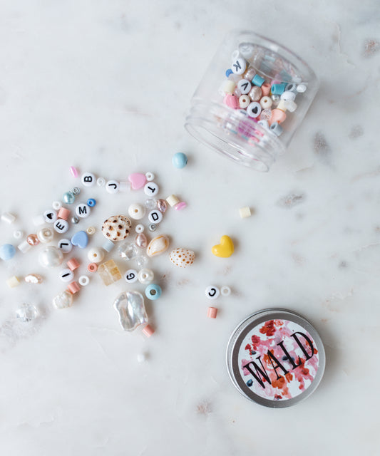A small jar tipped over, spilling assorted beads and letter charms onto a marble surface. A round tin labeled "NAIL'D" is placed nearby, perhaps part of the WALD World Candyman Kit in powder candy Tones.