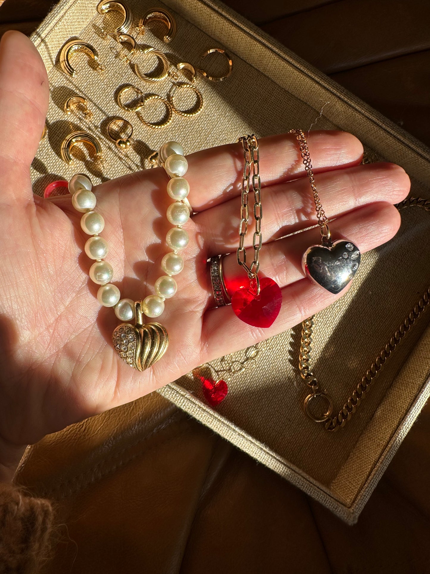 A hand displays a pearl bracelet with gold charms, earrings, a red heart pendant, and a silver ring. More jewelry rests in a beige tray beside the chic surprise WALD Berlin Bag by WALD Berlin.