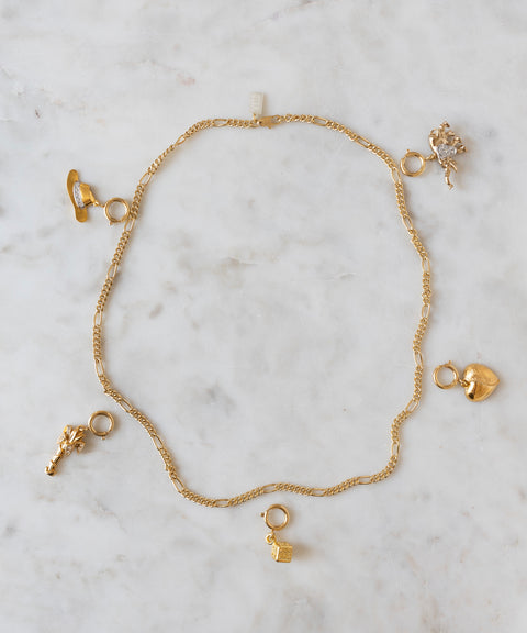 Showcased on a light marble surface, this gold chain necklace by WALD Berlin features five assorted gold charms—a shoe, lock, heart, animal, and floral shape—capturing the playful essence of the brand and offering a unique opportunity for personalization at the DIY Schmuck-Workshop x MINI im KaDeWe Berlin.