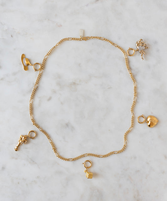 Auf einer hellen Marmorfläche präsentiert sich diese Goldkette von WALD Berlin mit fünf verschiedenen goldenen Anhängern – einem Schuh, einem Schloss, einem Herz, einem Tier und einer Blume –, die den verspielten Charakter der Marke einfangen und beim DIY-Schmuck-Workshop x MINI im KaDeWe Berlin eine einzigartige Gelegenheit zur individuellen Gestaltung bieten.
