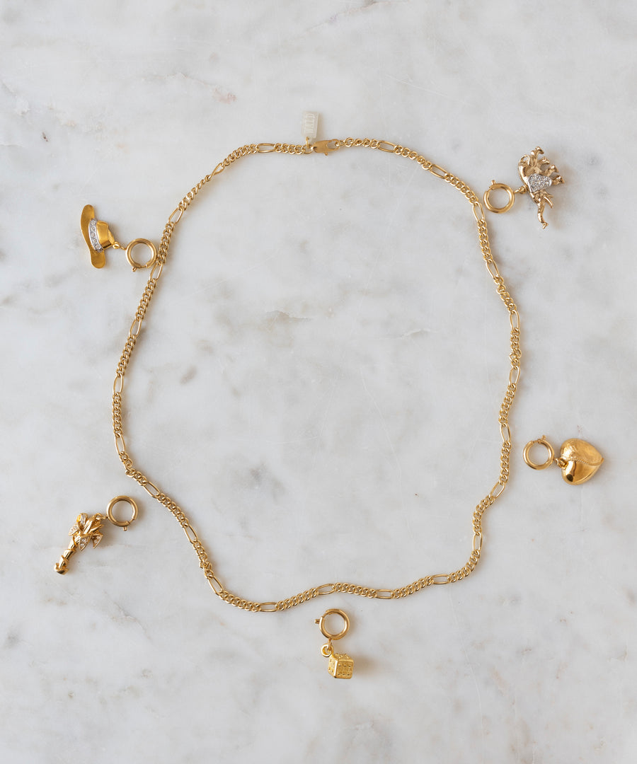 Showcased on a light marble surface, this gold chain necklace by WALD Berlin features five assorted gold charms—a shoe, lock, heart, animal, and floral shape—capturing the playful essence of the brand and offering a unique opportunity for personalization at the DIY Schmuck-Workshop x MINI im KaDeWe Berlin.