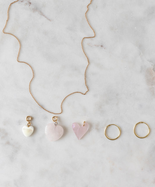 A Rose Quartz Heart Charm gold-plated brass necklace by WALD World, featuring three light rose, heart-shaped pendants, along with a pair of gold hoop earrings, are displayed on a marble surface.