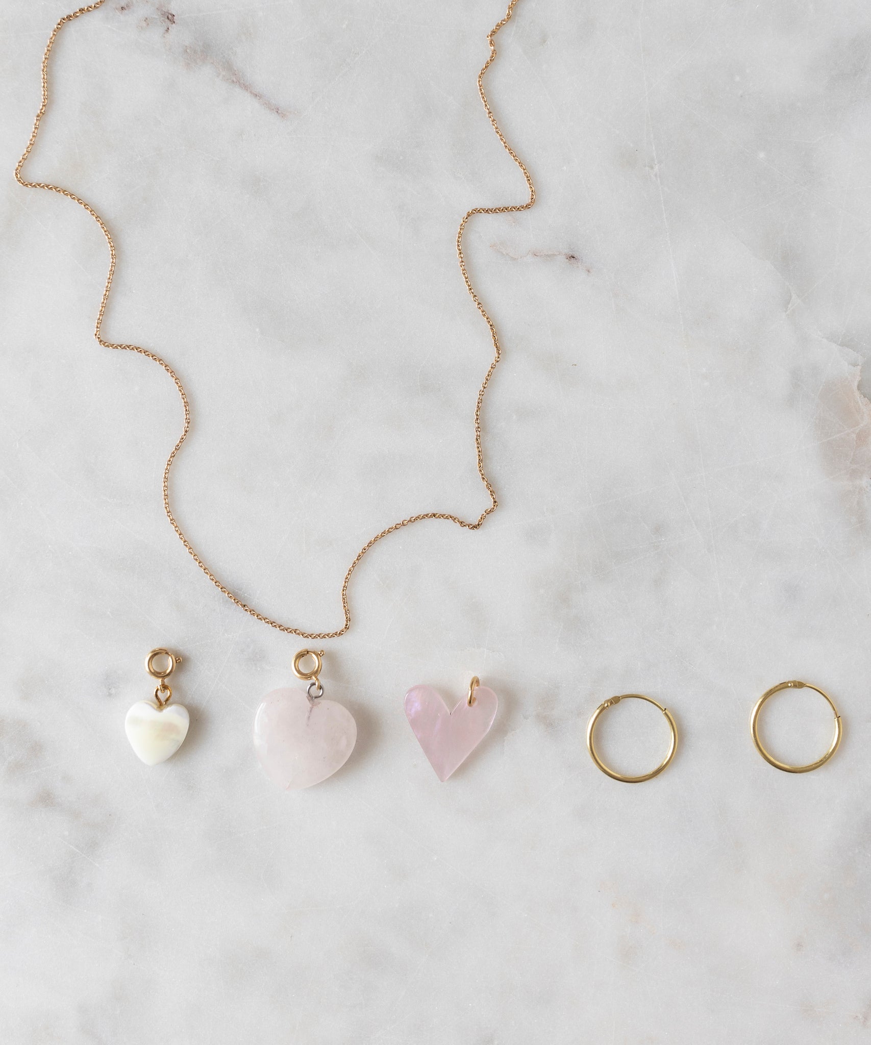 A Rose Quartz Heart Charm gold-plated brass necklace by WALD World, featuring three light rose, heart-shaped pendants, along with a pair of gold hoop earrings, are displayed on a marble surface.