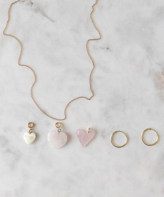 A Rose Quartz Heart Charm gold-plated brass necklace by WALD World, featuring three light rose, heart-shaped pendants, along with a pair of gold hoop earrings, are displayed on a marble surface.