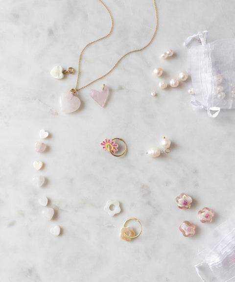 A Sweetwater Pearl Charm Set by WALD World, featuring heart-shaped designs and delicately crafted gold and silver necklaces, rings, and earrings adorned with sweetwater pearl charms and floral motifs, arranged on a white marble surface. A small mesh bag completes the collection.