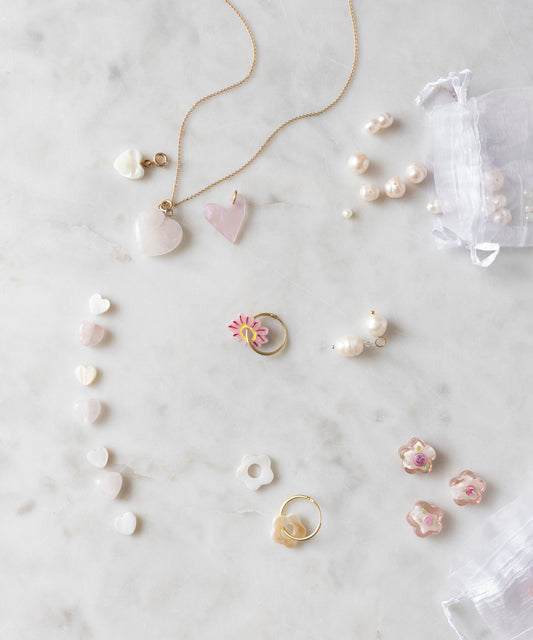 A Sweetwater Pearl Charm Set by WALD World, featuring heart-shaped designs and delicately crafted gold and silver necklaces, rings, and earrings adorned with sweetwater pearl charms and floral motifs, arranged on a white marble surface. A small mesh bag completes the collection.