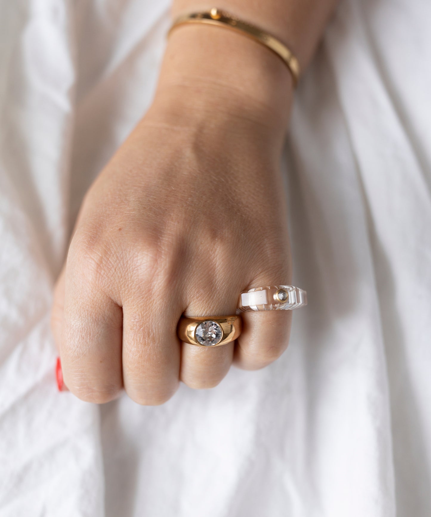 A hand with light skin wears an 18k gold-plated brass bracelet and WALD Berlin’s Pop Swarovski Ring—gold with a Swarovski Stone—plus a clear ring with a pearl, over white fabric. Made in Germany.