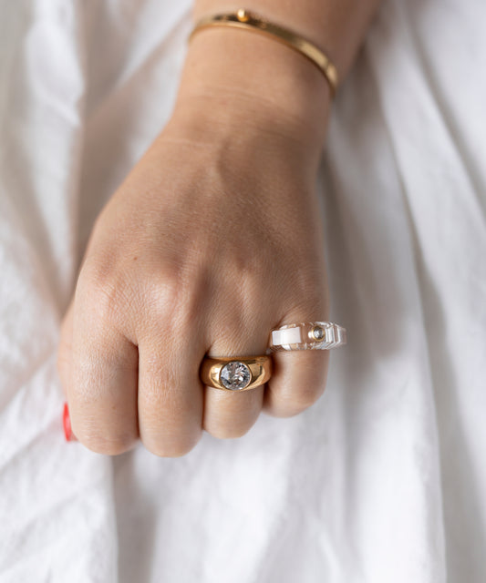 A hand with light skin wears an 18k gold-plated brass bracelet and WALD Berlin’s Pop Swarovski Ring—gold with a Swarovski Stone—plus a clear ring with a pearl, over white fabric. Made in Germany.